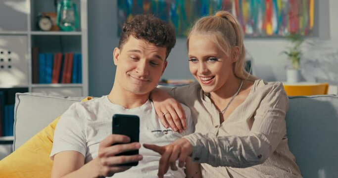 A couple of attractive teenagers in love are spending their free time after school together at home on couch, man with curly hair is holding phone, taking a picture with a smiling beautiful blonde