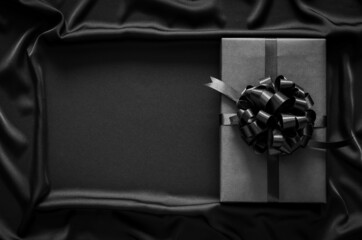 A black gift box with ribbon puts on black smooth and wavy cloth with space for text. Black friday and Boxing day concept.