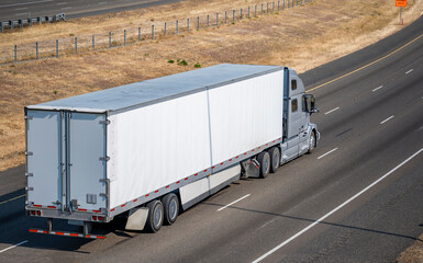Gray powerful industrial big rig semi truck transporting cargo in dry van semi trailer running on the summer divided highway road