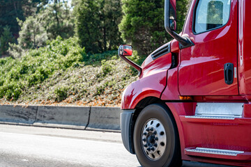 Side of the bright red big rig semi truck running on the road with forest on the hillside