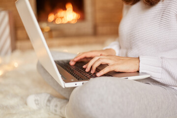 Faceless unknown woman sitting on floor on soft carpet at fireplace with laptop and working, typing, wearing white sweater and gray trousers, posing against burning fire on background.