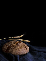 Round loaf of rye bread with a spike of wheat on a blue towel in a dark key on a black background, vertical photo, free space for text