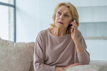 A sad elderly woman is talking on a mobile phone at home sitting on the couch