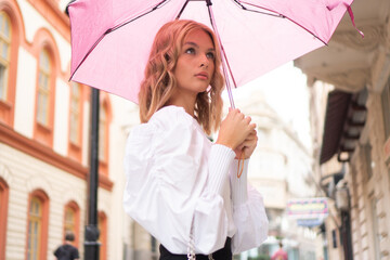 A portrait photo of a beautiful blond teenage girl with a pink umbrella on the street.