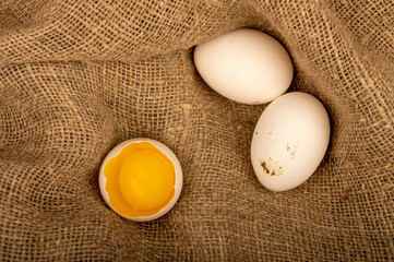 A broken white chicken egg and several whole eggs on a homespun fabric with a rough texture, close-up selective focus.