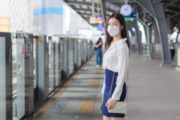 Obraz premium Asian professional business woman with long hair wears a mask is waiting to go to workplace or office by sky train in the New Normal lifestyle.