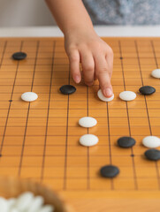 Child hand playing Go board game, white and black stones on the board game, wooden Asian classic game board with grid lines, Vertical image.