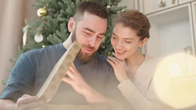 Chest-up of happy young Caucasian man opening box with Christmas gift from wife who hugging and looking at him smiling and talking, light coming out of box