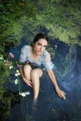 Fototapeta premium Art beautiful romantic woman lies in swamp in blue long dress with flowers. Portrait brunette in transparent dress in water swamp mud duckweed. Book cover