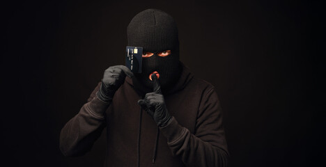 Thief in black balaclava holding black credit card. Banking and non cash thief concept - obrazy, fototapety, plakaty