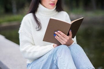 Obraz premium A young and attractive Caucasian brunette girl sitting and reading a book in a park near the river. The student is studying. The concept of education and knowledge.