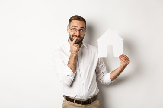 Real Estate. Man Thinking While Searching For Apartment, Holding Paper House Model, Standing Over White Background