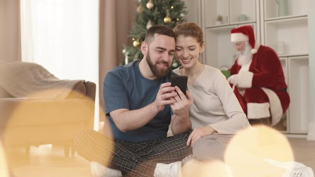 Locked-down of young Caucasian hetero couple sitting on floor in living room, using smartphone and smiling while sneaky Santa Claus putting presents under Christmas tree on background