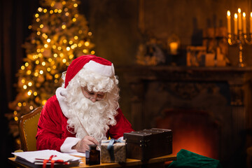 Obraz na płótnie Canvas Workplace of Santa Claus. Cheerful Santa is writing the letter while sitting at the table. Fireplace and Christmas Tree in the background. Christmas concept.
