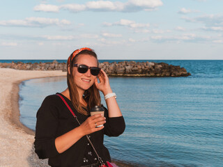 Pretty hipster woman in bandana long hair walking on sea background with coffee cup lifestyle real life travel photo.Millennial girl smiling drinking tea Relaxation Generation Z casual female portrait