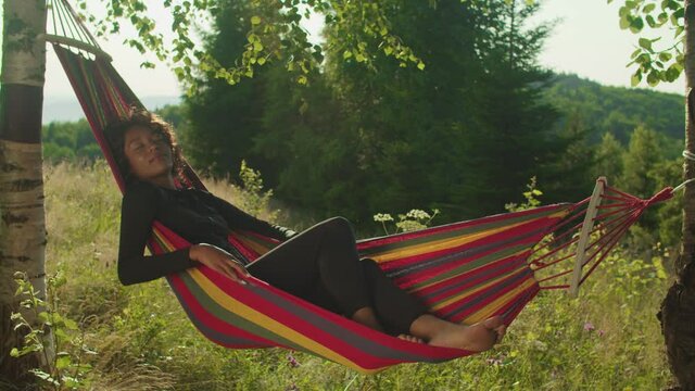 Serene pretty african female tourist sleeping in hammock on mountain hill, enjoying healthy lifestyle, freedom and recreation while relaxing on summer vacations in scenic mountain nature at dawn.
