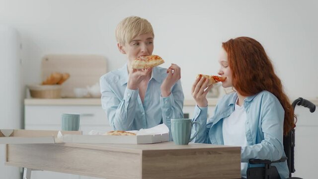 Little paralyzed girl in wheelchair having lunch with mother, eating tea and pizza, enjoying food delivery service