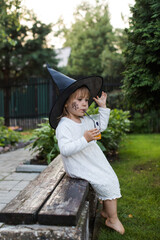 Little girl in dress and witch hat drinks juice in backyard