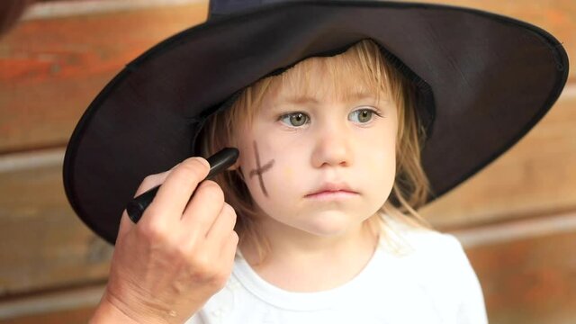 Little girl in black witch hat touches head by wooden house