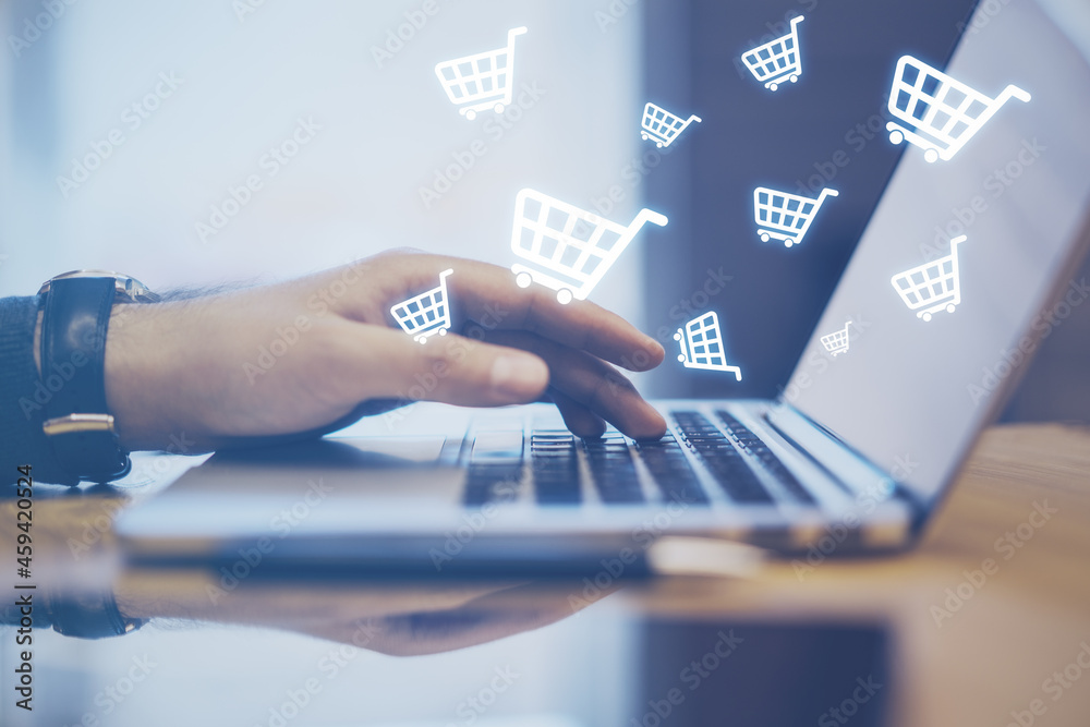 Poster Close up of hands using laptop computer with abstract glowing cart icons on blurry background. Online shopping, e-commerce and purchase concept.