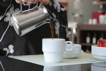 Woman's hand holding a coffee mug and pouring it into the coffee cup. Morning routine lifestyle concept, close up