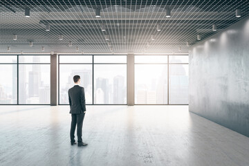 Attractive thoughtful young european businessman standing in empty interior with windows, city view and daylight.