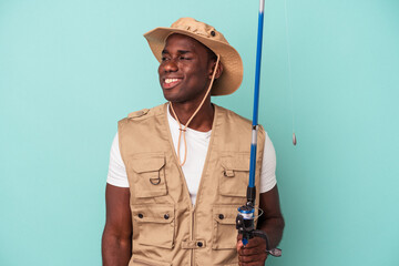 Young African American fisherman holding rod isolated on blue background looks aside smiling, cheerful and pleasant.