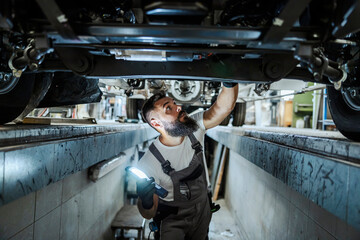 An auto-mechanic is standing in a car mechanic's pit and looking car bottom. He is lightning and looking for malfunction. A worker in the car repair workshop. - obrazy, fototapety, plakaty