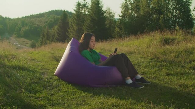 Charming asian woman traveler sitting on inflatable sofa, networking, text messaging and browsing social media content online while relaxing on mountain peak at daybreak during summer vacations.