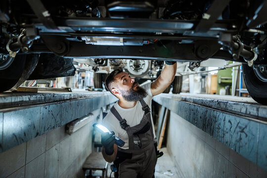 An auto-mechanic is standing in a car mechanic's pit and looking car bottom. He is lightning and looking for malfunction. A worker in the car repair workshop.
