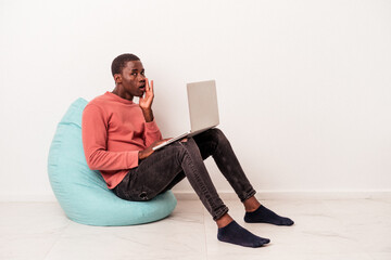 Young African American man sitting on a puff using laptop isolated on white background is saying a secret hot braking news and looking aside