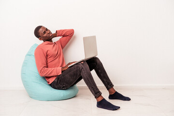 Young African American man sitting on a puff using laptop isolated on white background touching back of head, thinking and making a choice.