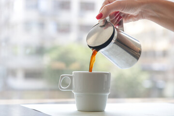 Woman's hand holding a coffee mug and pouring it into the coffee cup. Morning routine lifestyle concept, close up