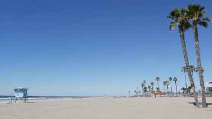 Tropical palm trees on white sandy beach by sea water waves, pacific ocean coast, Oceanside California USA. Blue sky and lifeguard tower. Life guard watchtower hut, summertime shore. Los Angeles vibes