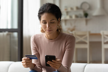 Young Caucasian woman relax at home shopping online on modern smartphone with credit debit card. Millennial female client or customer buy pay on web using secure banking system on cellphone.