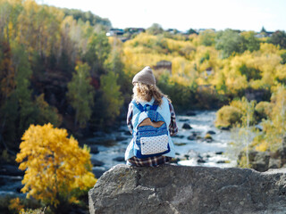 Young curly woman traveller in plaid t-shirt sitting on a rock; there is mountain river on a background; sustainable tourism; ecological tourism; discovery and explore