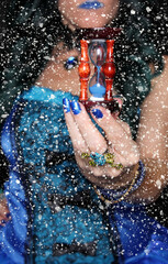 Fototapeta premium Woman in blue corset dress with hourglass in hand with snowflake bokeh