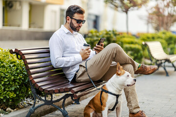 A middle-aged bearded businessman is sitting on a bench with his Stafford, drinking coffee to go and hanging on the social media on the phone.