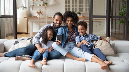 Happy African American family with two kids using smartphone, relaxing sitting on cozy couch, smiling mother and father with little son and daughter looking at phone screen, making video call