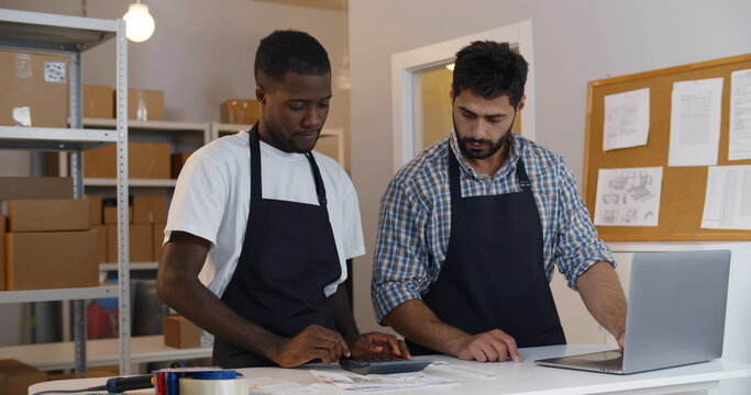 Small Business Owners Count Profit Using Laptop And Calculator In Warehouse