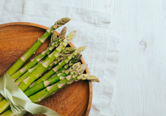 A bouquet of green fresh asparagus is tied with a green silk ribbon. Asparagus lies on a round wooden plate on a light table with a linen napkin, top view. Copy space.