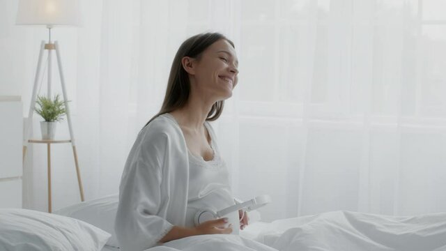 Pregnant Woman Holding Headphones Near Belly And Dancing While Sitting In Bed