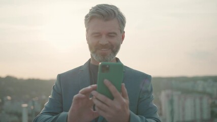 Bearded lucky smiling caucasian adult businessman on rooftop, holding smartphone and texting messages. People, success and career. Outdoor background. - Powered by Adobe