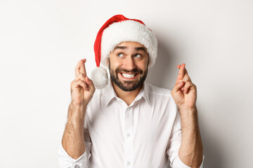 Party, winter holidays and celebration concept. Happy man in santa hat making christmas wish, cross fingers for good luck and smiling excited, white background - Powered by Adobe