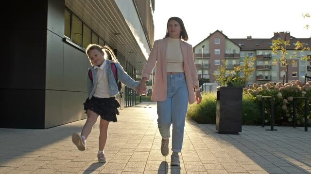Young woman leads her daughter 6-7 years old to school by the hand. The girl with pigtails jumps happily on the go. Back to school.