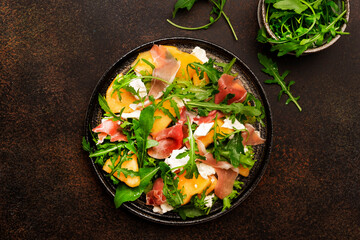 Salad with melon, jamon, goat cheese and arugula on brown background, top view, copy space