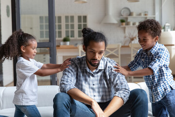 Unhappy exhausted African American father ignoring noisy children, feeling exhausted, sitting on couch at home, little son and daughter demanding tired depressed dad attention, fatherhood concept