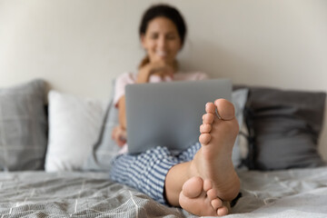 Crop close up of young woman sit relax at home on bed use laptop gadget. Closeup of female legs...