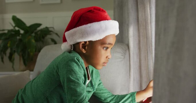 Animation of sad african american boy wearing santa hat looking outside window at christmas time