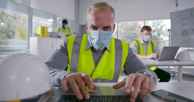 Mature architect in face mask working on laptop sitting at table in office